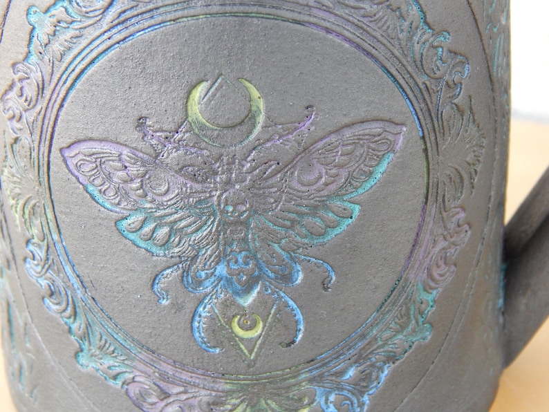 May include: A dark gray ceramic mug with a detailed embossed design. The central image is a moth with a crescent moon and geometric shapes, framed by an ornate border. The moth and border have iridescent blue, green, and purple accents.