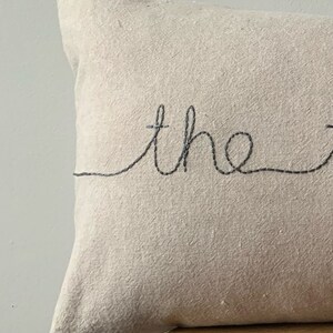 May include: A beige linen pillow with the word "the" embroidered in black thread.