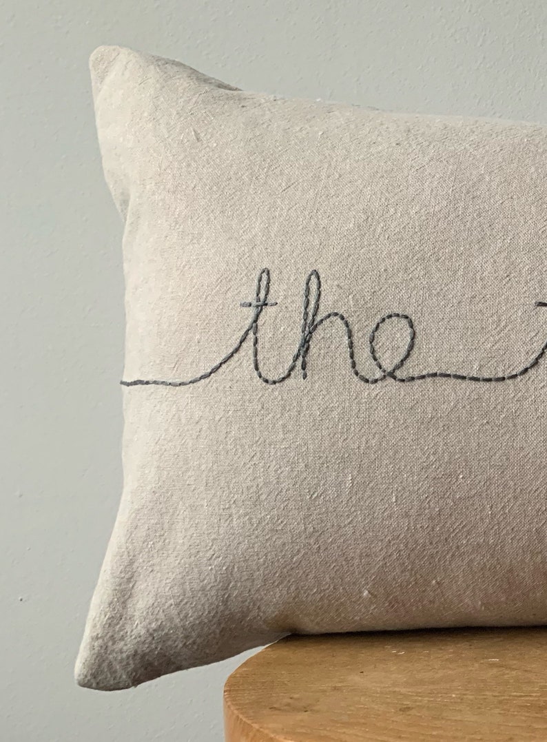 May include: A beige linen pillow with the word "the" embroidered in black thread.