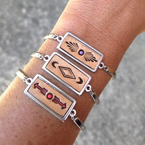 May include: Three silver bracelets with rectangular, tan leather pendants. Each pendant features a different design: a sunburst with a purple gem, a crescent moon and diamond, and an arrow. The bracelets are worn on a wrist.