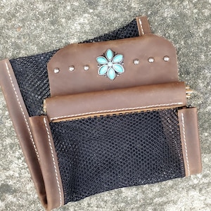 May include: A brown leather saddle bag with a black mesh pocket and a turquoise flower design. The bag has a zipper closure and is decorated with metal studs.