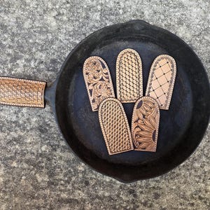 May include: A collection of intricately carved leather pot holders and a matching handle cover, displayed in a black cast iron skillet. The pot holders feature various embossed patterns, including floral and geometric designs. The leather is a light brown color.