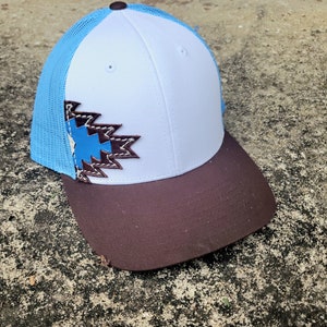 May include: A white and blue trucker hat with a brown brim. The hat has a blue and brown geometric design on the front.