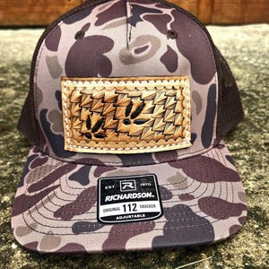 May include: A camouflage trucker hat with a brown mesh back and a leather patch. The patch features a woven design with a leaf pattern. The hat has a Richardson 112 Trucker label.
