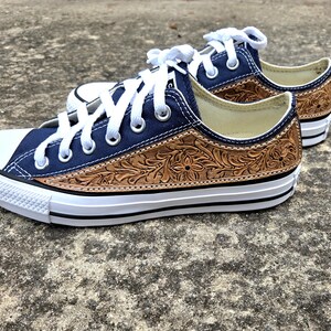 May include: A pair of navy blue canvas sneakers with white laces and a white rubber toe cap. The sides feature a brown, intricately carved leather design. The shoes have a white sole with a black stripe.