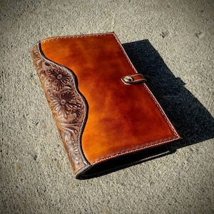 May include: A handcrafted leather journal with a rich, warm brown color. The cover features a floral embossed design on the left side and a smooth, lighter brown surface on the right. A gold-colored clasp secures the journal.
