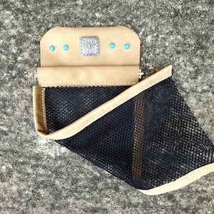 May include: A tan leather pouch with a black mesh pocket. The pouch has a silver decorative square in the center and three turquoise-colored stones. The edges are stitched with a light tan thread.
