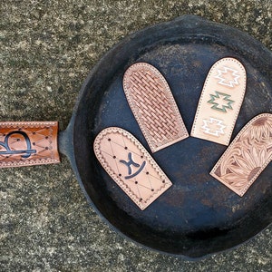 May include: A collection of leather cast iron skillet handle covers in various designs, including a woven pattern, geometric shapes, and a sunflower. The covers are in shades of brown and tan, with black accents. The handle cover on the left has a black letter "B".
