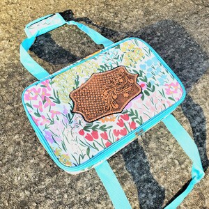 May include: A floral print zippered tote bag with a brown leather patch featuring a decorative design. The bag has two turquoise straps.