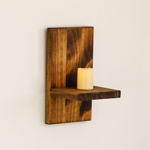 May include: A rustic, wooden wall-mounted candle holder. The holder is stained a warm brown and features a small shelf to hold a single candle. A cream-colored candle sits on the shelf, ready to be lit. The design is simple and elegant.
