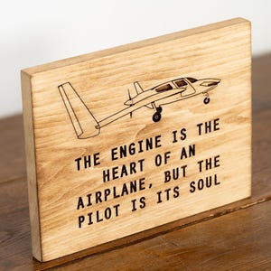 May include: Wooden plaque with a laser-etched airplane design and the quote: "THE ENGINE IS THE HEART OF AN AIRPLANE, BUT THE PILOT IS ITS SOUL." The plaque is made of light-colored wood.
