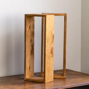 May include: Two rectangular wooden decorative boxes with open sides. One box is upright, and the other is leaning against it. The wood has a natural, light brown color with visible grain and knots. They sit on a dark wooden table.