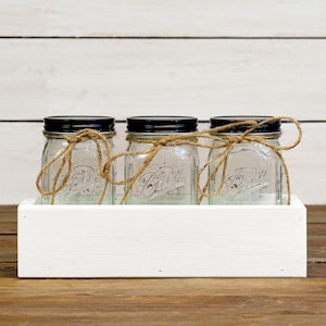 May include: Three clear glass jars with black lids and twine tied around the top are displayed in a white wooden box. The jars are sitting on a wooden surface.