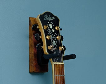 May include: A black and gold electric guitar with the word "Hofner" on the headstock is mounted on a wooden wall hanger.