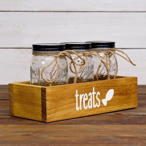 May include: A wooden box with three clear glass jars with black lids. The jars are tied with twine and sit inside the box. The box has the word "treats" and a bone-shaped graphic in white.