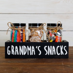 May include: Three glass jars with black lids and twine tied around the lids, filled with different snacks, are displayed in a black wooden box with the text "Grandma's Snacks" written in white.