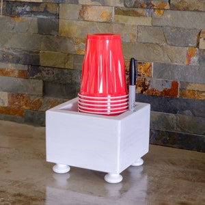 May include: White wooden cup holder with four legs, holding a stack of red plastic cups and a black marker.