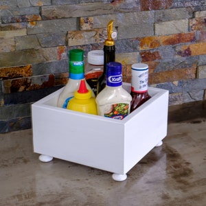 May include: White wooden condiment holder with four legs. The holder contains five bottles of various sauces and dressings, including a bottle of Kraft salad dressing.