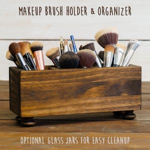 May include: A wooden makeup brush holder and organizer with a dark stain. The rectangular box is filled with various makeup brushes. The text "MAKEUP BRUSH HOLDER & ORGANIZER" is at the top, and "OPTIONAL GLASS JARS FOR EASY CLEANUP" is at the bottom.