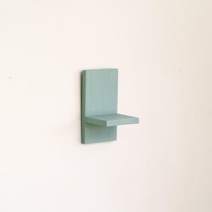 May include: A small, wall-mounted shelf painted in a muted teal color. The shelf has a rectangular back panel and a horizontal shelf extending from it. The design is minimalist and modern, suitable for displaying small items.
