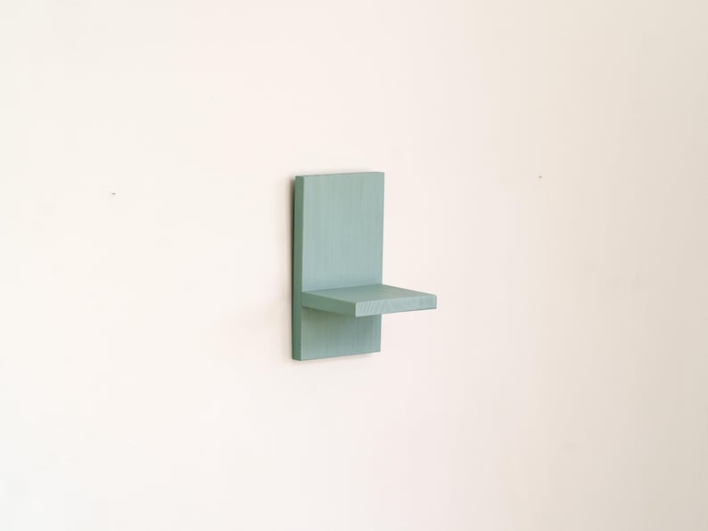 May include: A small, wall-mounted shelf painted in a muted teal color. The shelf has a rectangular back panel and a horizontal shelf extending from it. The design is minimalist and modern, suitable for displaying small items.