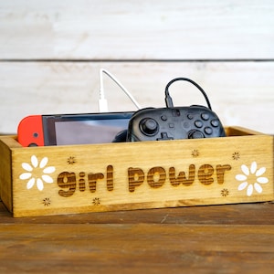 May include: A wooden box with the words "girl power" engraved on the front. The box contains a video game console and a controller. The box is decorated with white daisy flowers.