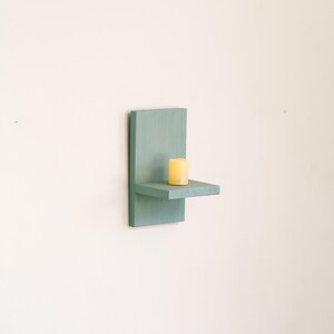 May include: A wall-mounted teal wooden shelf with a small, yellow candle. The shelf has a rectangular back and a small, horizontal platform. The candle is cylindrical. The shelf is a decorative item.