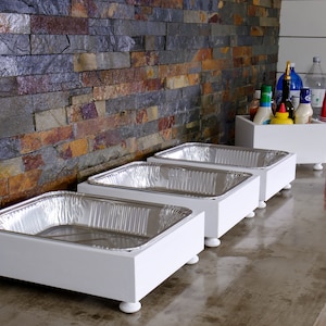 May include: Four white wooden serving trays with silver aluminum food pans on top. The trays have four small white feet and are lined up on a concrete countertop.