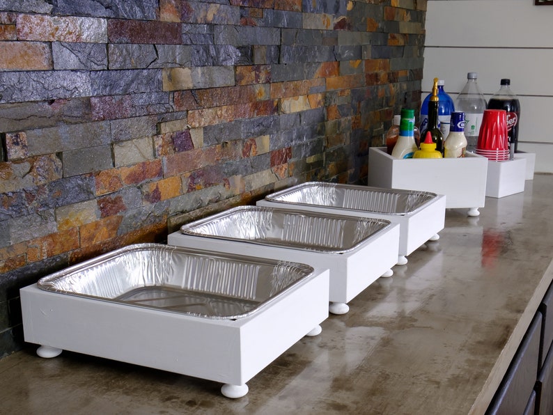 May include: Four white wooden serving trays with silver aluminum food pans on top. The trays have four small white feet and are lined up on a concrete countertop.