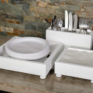 May include: White wooden countertop organiser with three separate compartments. The left compartment holds a stack of white paper plates. The middle compartment holds a set of cutlery. The right compartment holds a stack of white napkins.