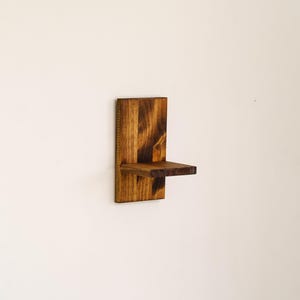 May include: A small, wall-mounted wooden shelf with a dark brown finish. The shelf has a rectangular back piece and a small, horizontal shelf extending outward. The wood grain is visible, adding texture to the design. The shelf is designed to hold small items.