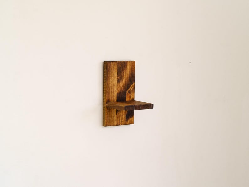 May include: A small, wall-mounted wooden shelf with a dark brown finish. The shelf has a rectangular back piece and a small, horizontal shelf extending outward. The wood grain is visible, adding texture to the design. The shelf is designed to hold small items.