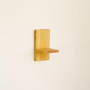May include: A small, wall-mounted wooden shelf. The shelf is made of light-colored wood and has a simple, minimalist design. The shelf is rectangular with a small ledge for displaying items. The shelf is attached to a vertical wooden backboard.