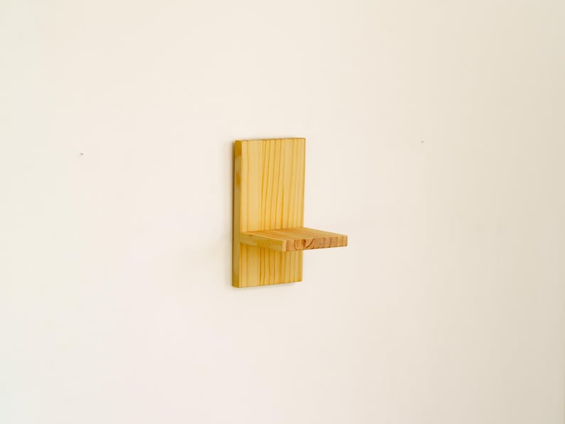 May include: A small, wall-mounted wooden shelf. The shelf is made of light-colored wood and has a simple, minimalist design. The shelf is rectangular with a small ledge for displaying items. The shelf is attached to a vertical wooden backboard.