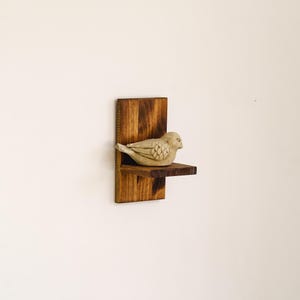 May include: A small, decorative wooden shelf with a bird figurine. The shelf is made of dark stained wood and has a simple, minimalist design. The bird is a light beige color and is perched on the shelf. The shelf is mounted on a white wall.