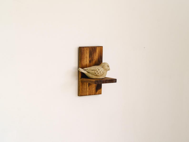 May include: A small, decorative wooden shelf with a bird figurine. The shelf is made of dark stained wood and has a simple, minimalist design. The bird is a light beige color and is perched on the shelf. The shelf is mounted on a white wall.