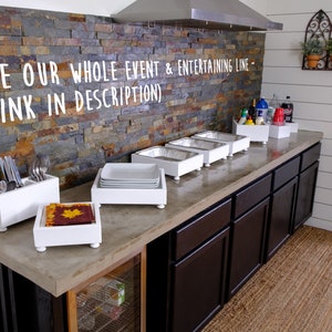 May include: White wooden serving trays with metal containers on a concrete countertop. The trays are set up for a buffet or party. The countertop is in front of a stone wall and dark brown cabinets.  See our whole event & entertaining line - (link in description)