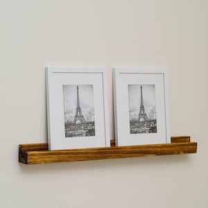 May include: Two white picture frames with black and white photos of the Eiffel Tower in Paris, France, are displayed on a dark brown wooden shelf.