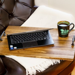 May include: A wooden laptop tray with black handles, holding a black laptop and a black mug with green text that reads "Williamsburg, VA". The tray is placed on a brown leather armchair with a white blanket.
