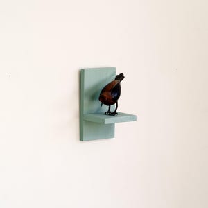 May include: A decorative bird sculpture with a brown body and black legs stands on a small, light blue shelf. The shelf is mounted on a matching rectangular backing, creating a minimalist display against a white wall.