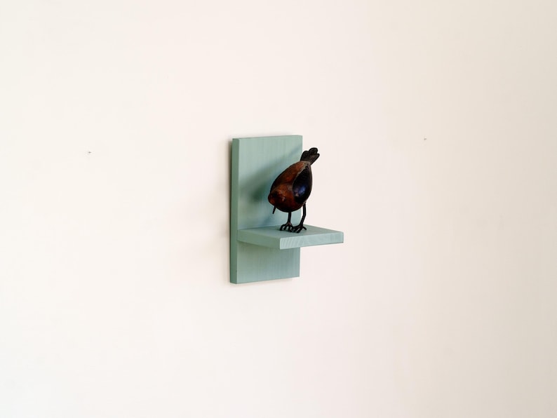 May include: A decorative bird sculpture with a brown body and black legs stands on a small, light blue shelf. The shelf is mounted on a matching rectangular backing, creating a minimalist display against a white wall.