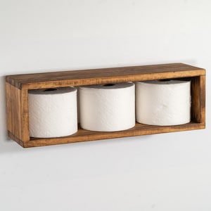 May include: A wooden bathroom shelf with three rolls of white toilet paper. The shelf is rectangular, stained a dark brown, and mounted on a white wall. The rolls are evenly spaced within the shelf's compartments.