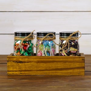 May include: Three glass jars with black lids and twine tied around the tops, filled with different types of candy. The jars are sitting in a wooden crate.