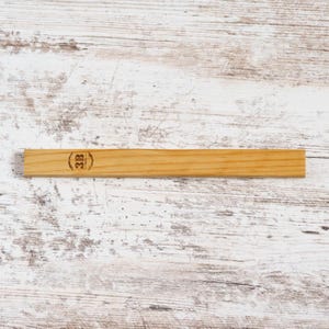 Peut inclure: Un objet rectangulaire en bois de couleur marron clair. Il comporte un tampon circulaire avec les lettres "3B". Le grain du bois est visible. L'objet repose sur une surface en bois blanc et marron clair. Il pourrait s'agir d'une règle.