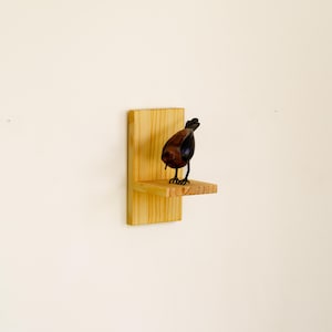 May include: A decorative bird figurine with a brown body and black accents stands on a small wooden shelf. The shelf is attached to a larger wooden backing, creating a rustic display. The bird is perched on the shelf, adding a touch of nature to the decor.