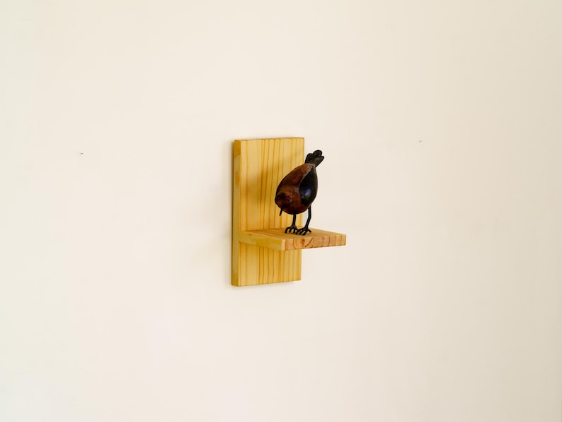 May include: A decorative bird figurine with a brown body and black accents stands on a small wooden shelf. The shelf is attached to a larger wooden backing, creating a rustic display. The bird is perched on the shelf, adding a touch of nature to the decor.