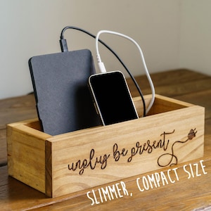 May include: Wooden charging station with a phone and a tablet inside. The station has the words "unplug be present" and "SLIMMER, COMPACT SIZE" engraved on the front. Black and white charging cables are connected to the devices.