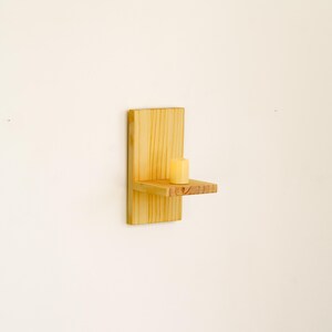 May include: A small, wall-mounted wooden shelf with a single candle. The shelf is made of light-colored wood and has a simple, minimalist design. The candle is a pale yellow color and is centered on the shelf.