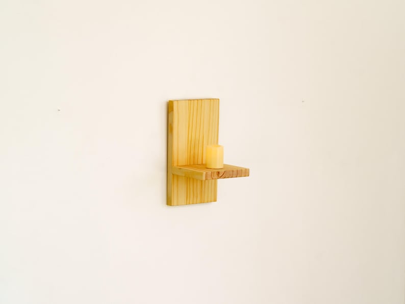 May include: A small, wall-mounted wooden shelf with a single candle. The shelf is made of light-colored wood and has a simple, minimalist design. The candle is a pale yellow color and is centered on the shelf.