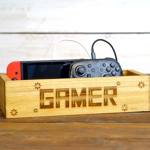 May include: A wooden box with the word "GAMER" engraved on the front. The box is holding a video game console and a controller.